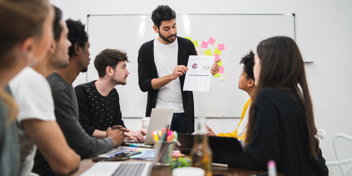 A man stands before a group seated at a table, discussing lead generation services in a collaborative setting.