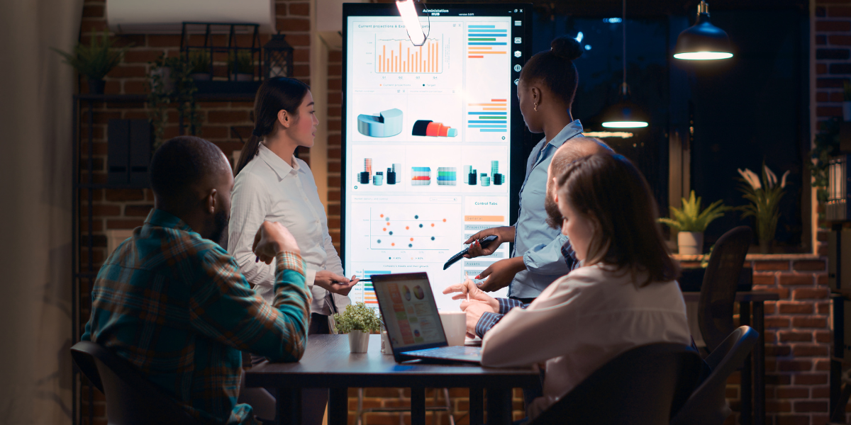 People seated at a table, engaged in discussion, with a large screen displaying analytics data in the background.